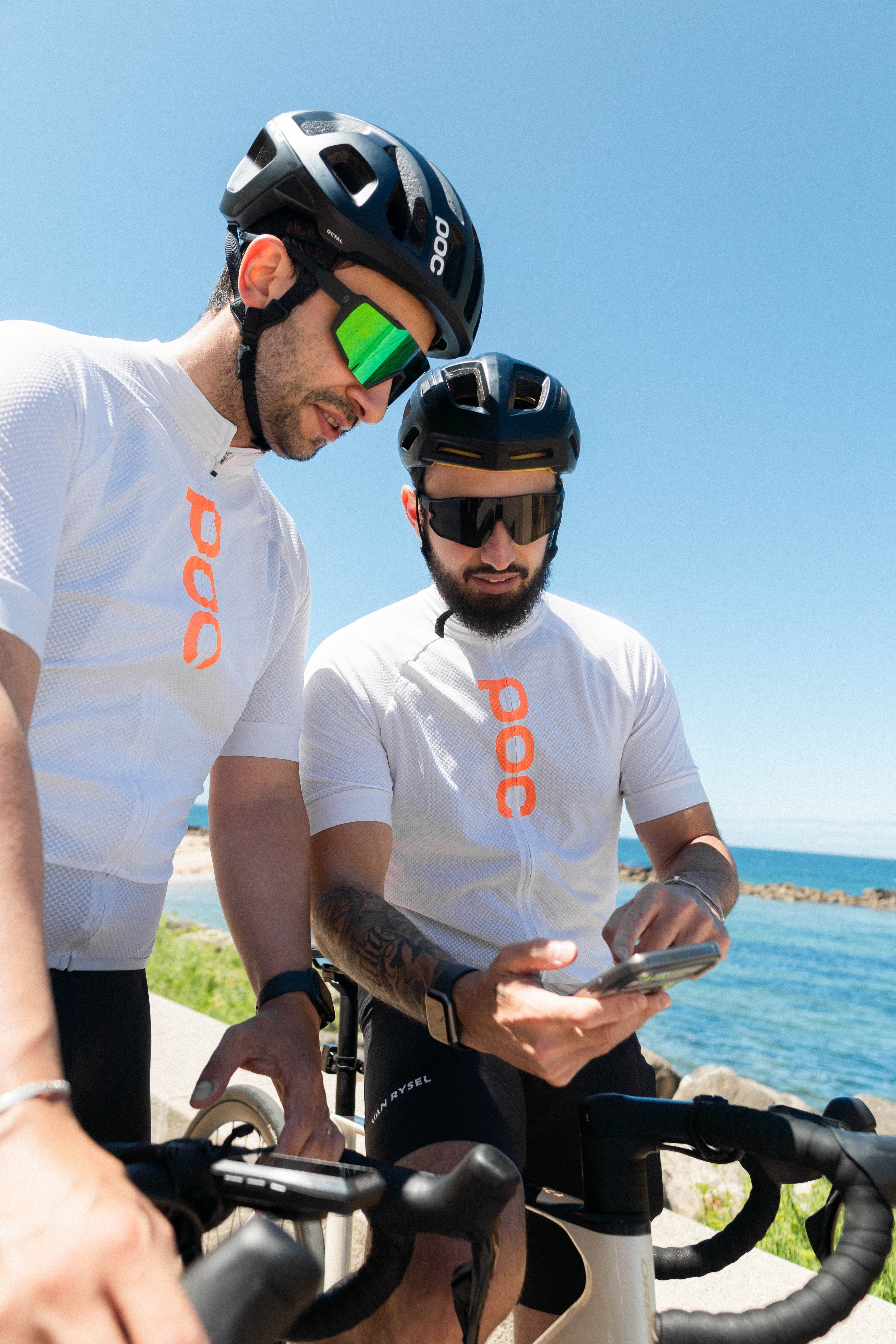 Two cyclists paused by the sea, one checking a training app on a phone
